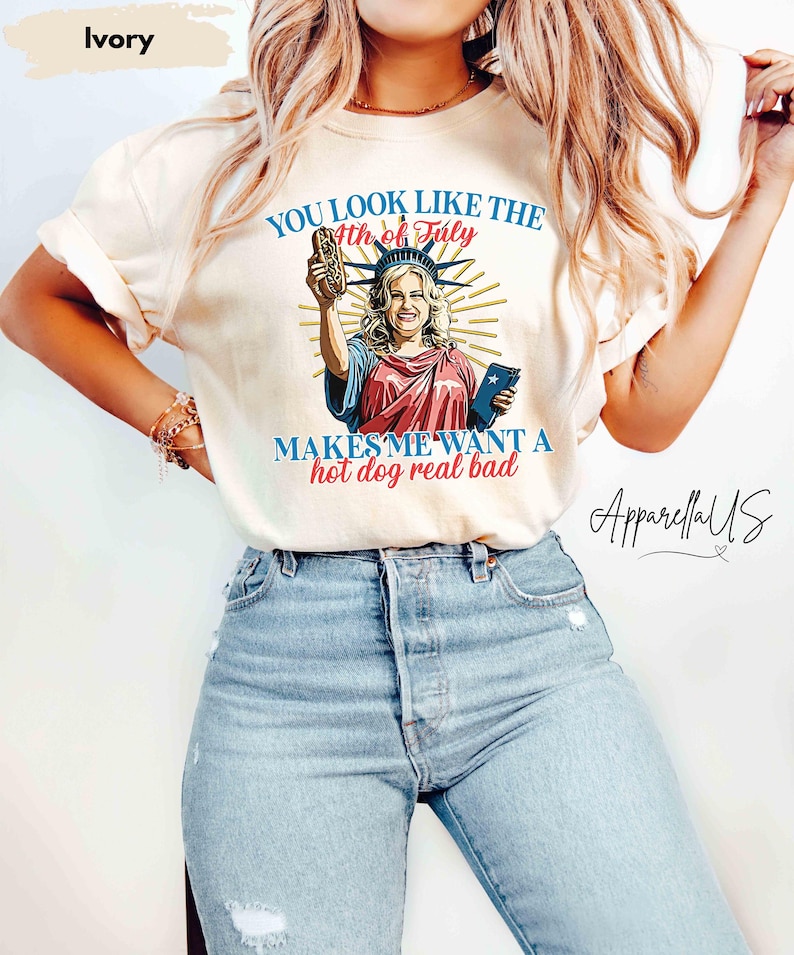 Puede incluir: Camiseta de color marfil con un gr&aacute;fico de la Estatua de la Libertad sosteniendo un perrito caliente. El texto dice "YOU LOOK LIKE THE 4th of July MAKES ME WANT A hot dog real bad" en azul y rojo. La marca "ApparellaUS" est&aacute; en la esquina inferior derecha.