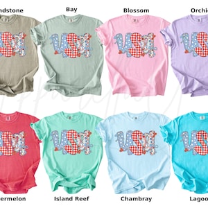 May include: A collection of eight short-sleeved t-shirts in various colors, each featuring the word "USA" in a patriotic design. The letters are a mix of blue polka dots and red gingham patterns. The shirts are labeled with color names.