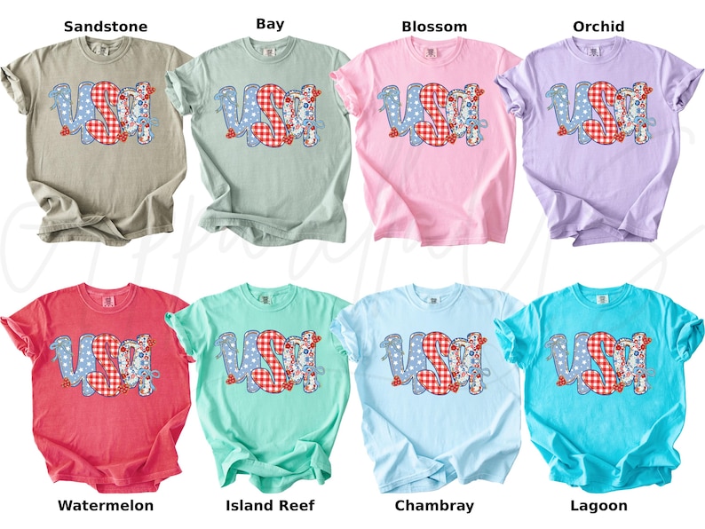 May include: A collection of eight short-sleeved t-shirts in various colors, each featuring the word "USA" in a patriotic design. The letters are a mix of blue polka dots and red gingham patterns. The shirts are labeled with color names.