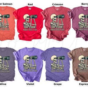 Op de afbeelding: Een verzameling van acht T-shirts in verschillende kleuren, elk met een skelet dat door een verrekijker kijkt. De tekst "OH LOOK! NOBODY GIVES A SHIT" staat onder het skelet. Kleuren zijn helder zalm, rood, karmozijn, bes, wijn, violet, druif en espresso.