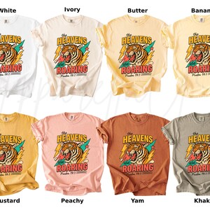 May include: A collection of t-shirts in various colors, including white, ivory, butter, banana, mustard, peachy, yam, and khaki. Each shirt features a graphic of a tiger with the text "The Heavens are Roaring" and "Psalm 19:1".