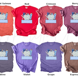 May include: A set of ten t-shirts featuring a colorful illustration of a patterned apple with school supplies. The shirts are available in ten different colors: bright salmon, red, crimson, berry, wine, violet, grape, espresso, teal, and navy.