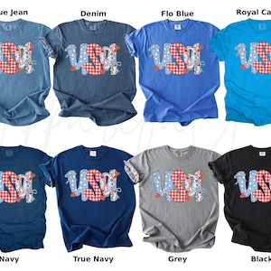 May include: A collection of eight t-shirts in various colors, including blue, navy, grey, and black. Each shirt features the word "USA" in a patriotic design with red, white, and blue elements. The shirts are short-sleeved.