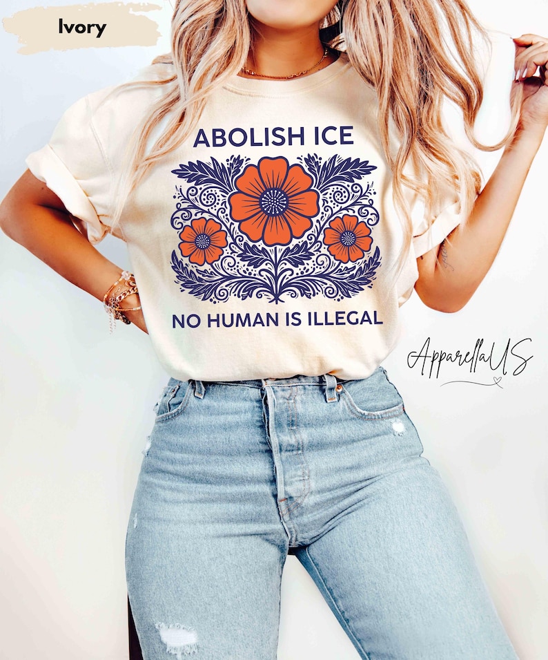May include: Ivory-colored t-shirt with the text "ABOLISH ICE" and "NO HUMAN IS ILLEGAL" in blue. The shirt features a floral design with orange flowers and blue leaves. The shirt is paired with light blue jeans.