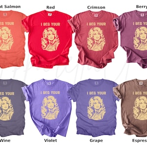 May include: A collection of eight short-sleeve t-shirts in various colors including bright salmon, red, crimson, berry, wine, violet, grape, and espresso. Each shirt features a graphic of a woman with wavy hair and the text I BEG YOUR.