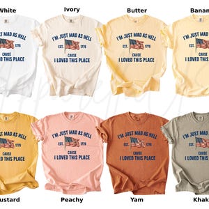 May include: A collection of short-sleeved t-shirts in various colors, including white, ivory, and mustard. Each shirt features the text "I'M JUST MAD AS HELL CAUSE I LOVED THIS PLACE" with an American flag graphic.