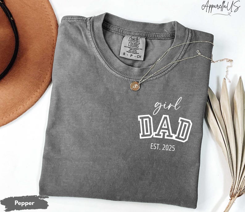 May include: A gray Comfort Colors t-shirt with the words "girl DAD EST. 2025" in white. A gold necklace with a pendant is draped on the shirt. A brown hat is visible in the upper left corner.