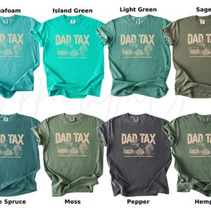 May include: A collection of eight t-shirts in various colors, including seafoam, island green, light green, sage, blue spruce, moss, pepper, and hemp. Each shirt features the text "DAD TAX" in a distressed font, along with a graphic. The shirts are short-sleeved.