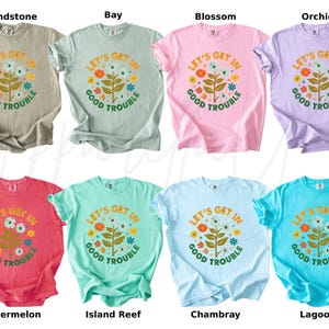 Peut inclure: Huit t-shirts de couleurs vari&eacute;es, chacun portant l'inscription "LET'S GET IN GOOD TROUBLE" dans un motif floral. Les couleurs comprennent le beige, le vert clair, le rose, le violet, le rouge, le turquoise, le bleu clair et le bleu sarcelle.