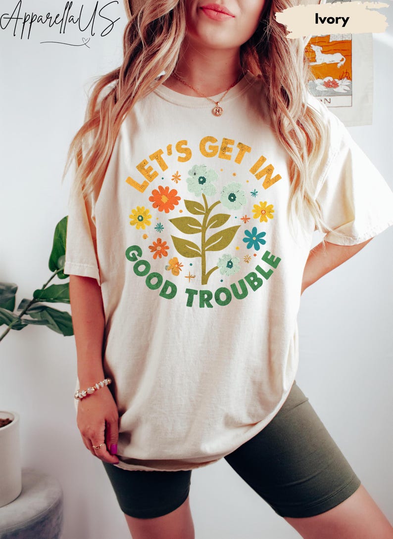 Peut inclure: T-shirt ivoire avec un motif floral et le texte "LET'S GET IN GOOD TROUBLE" en orange et vert. Le t-shirt pr&eacute;sente un motif floral rond avec des fleurs et des feuilles color&eacute;es. C'est un v&ecirc;tement d&eacute;contract&eacute; pour tous les jours.