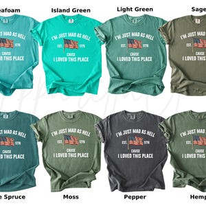 May include: A collection of eight t-shirts in various colors, including seafoam, island green, light green, sage, blue spruce, moss, pepper, and hemp. Each shirt features the text "I'M JUST MAD AS HELL" with an American flag graphic and the text "CAUSE I LOVED THIS PLACE."
