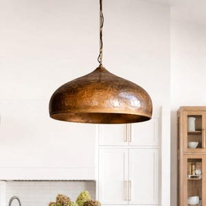Op de afbeelding: Een keukeninterieur met een grote, koperkleurige hanglamp boven een wit aanrecht eiland. Twee houten barkrukken met geweven zittingen staan voor het eiland. Een vaas met gedroogde bloemen staat op het aanrecht.