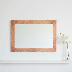 May include: A rectangular mirror with a hammered copper frame. The mirror is displayed on a white wall with a white shelf below. A clear vase with white flowers is to the right of the mirror.