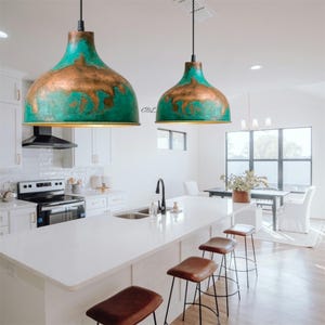 Puede incluir: Dos l&aacute;mparas colgantes con pantallas de color turquesa y cobre cuelgan sobre una isla de cocina blanca. La cocina cuenta con una encimera blanca, electrodom&eacute;sticos de acero inoxidable y taburetes de bar de cuero marr&oacute;n.