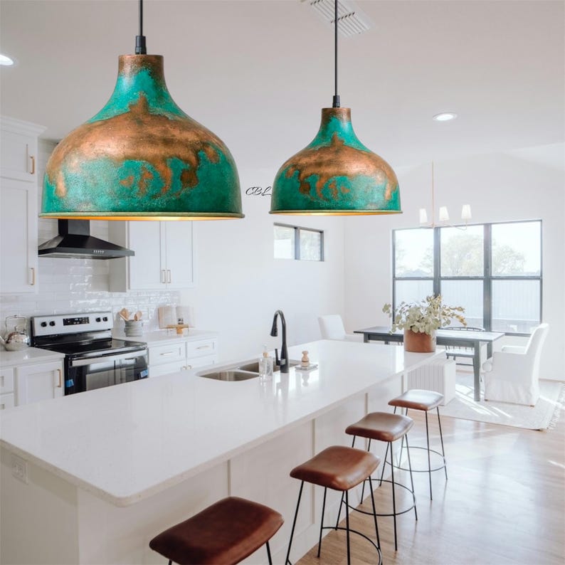 Puede incluir: Dos l&aacute;mparas colgantes con pantallas de color turquesa y cobre cuelgan sobre una isla de cocina blanca. La cocina cuenta con una encimera blanca, electrodom&eacute;sticos de acero inoxidable y taburetes de bar de cuero marr&oacute;n.