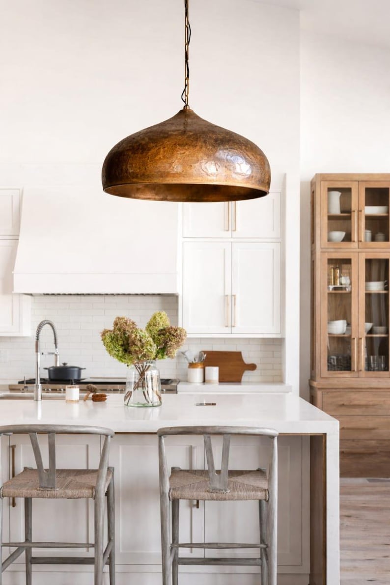 Op de afbeelding: Een keukeninterieur met een grote, koperkleurige hanglamp boven een wit aanrecht eiland. Twee houten barkrukken met geweven zittingen staan voor het eiland. Een vaas met gedroogde bloemen staat op het aanrecht.