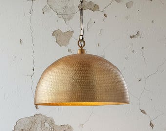 Puede incluir: Una lámpara colgante dorada en forma de cúpula con una textura martillada. La luz cuelga de una cadena y un pequeño accesorio circular. El fondo es una pared blanca desgastada con grietas y pintura descascarillada.