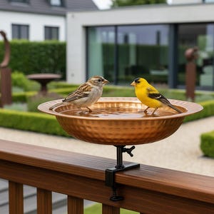May include: A copper-colored bird bath with a textured surface, mounted on a black clamp. Two birds, one brown and one yellow, are perched on the edge of the bath, which is filled with water. The bird bath is attached to a wooden railing.