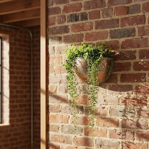 Puede incluir: Una maceta de pared en forma de media luna de color cobre llena de plantas verdes colgantes, montada en una pared de ladrillo rojo. La maceta tiene un diseño floral en relieve. La pared de ladrillo está iluminada por la luz del sol.
