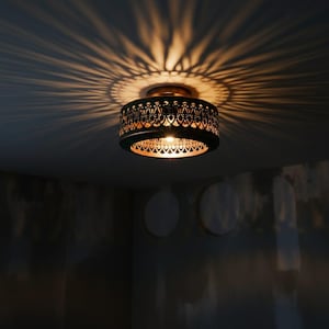 May include: A decorative ceiling light fixture with a dark metal shade featuring an intricate, openwork design. The light casts a patterned shadow on the ceiling. The interior of the shade has a warm, golden hue.