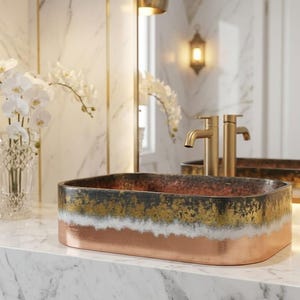 Op de afbeelding: Rechthoekige wastafel met een koperen, gouden en zwarte afwerking. De wastafel staat op een marmeren aanrecht, met gouden kranen en een spiegel op de achtergrond. Een vaas met witte orchideeën voegt elegantie toe.