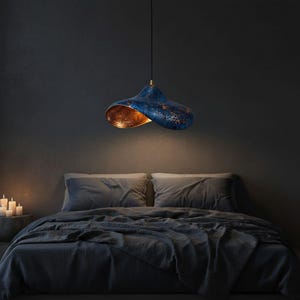 May include: A modern, blue and gold pendant light fixture hangs above a bed. The light has a unique, curved shape with a textured blue exterior and a warm gold interior. The bedroom has a dark, moody aesthetic with gray bedding.