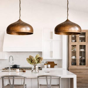 Op de afbeelding: Twee koperkleurige hanglampen hangen boven een wit keukeneiland. De lampen hebben een gehamerde textuur en een afgeronde, klokvormige vorm. De keuken heeft witte kasten, een wit aanrechtblad en een houten kast met glazen deuren.