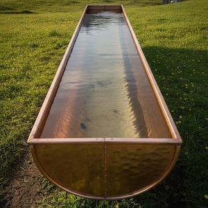 Puede incluir: Un largo abrevadero rectangular de cobre lleno de agua, colocado sobre hierba verde. El abrevadero tiene una textura martillada en los lados y una superficie lisa y reflectante en el interior. El agua refleja el cielo y el entorno.