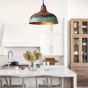 Puede incluir: Una l&aacute;mpara colgante de cobre y verde azulado cuelga sobre una isla de cocina con dos sillas tejidas. La l&aacute;mpara tiene forma de c&uacute;pula con acabado bronce y borde verde azulado. La cocina cuenta con gabinetes blancos y un gabinete de madera.