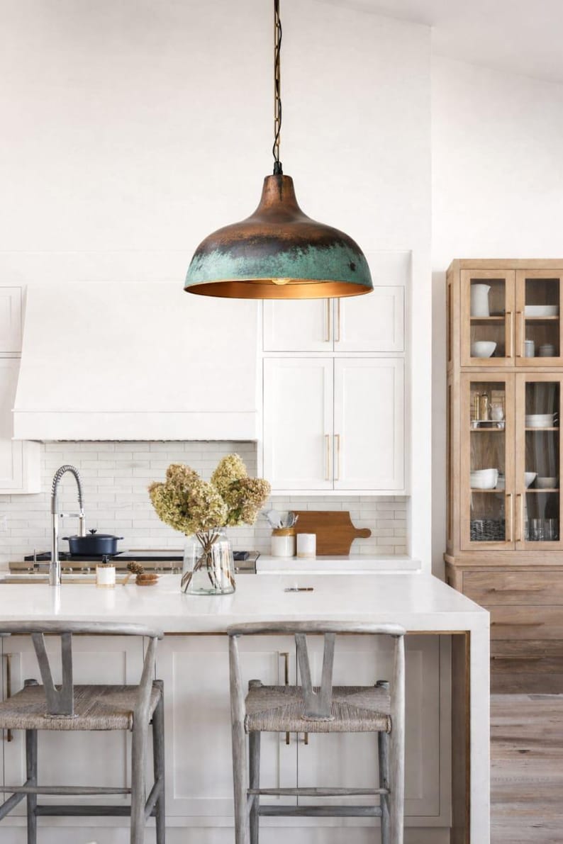 Puede incluir: Una l&aacute;mpara colgante de cobre y verde azulado cuelga sobre una isla de cocina con dos sillas tejidas. La l&aacute;mpara tiene forma de c&uacute;pula con acabado bronce y borde verde azulado. La cocina cuenta con gabinetes blancos y un gabinete de madera.