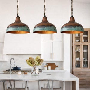 Puede incluir: Tres l&aacute;mparas colgantes con acabado cobrizo y turquesa cuelgan sobre una isla de cocina. Las l&aacute;mparas tienen forma de c&uacute;pula y est&aacute;n suspendidas por cadenas. La cocina cuenta con gabinetes blancos y una encimera de color claro.