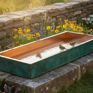 Puede incluir: Un abrevadero rectangular de cobre y verdín lleno de agua, apoyado sobre una superficie de piedra. Varios pájaros pequeños se posan en el borde. El fondo presenta un muro de piedra, flores y vegetación.