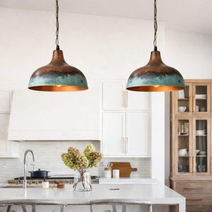 Puede incluir: Dos l&aacute;mparas colgantes con pantallas de color cobre y una banda turquesa. Las luces cuelgan de cadenas negras en un entorno de cocina. La cocina cuenta con gabinetes blancos, una encimera blanca y un jarr&oacute;n con flores.
