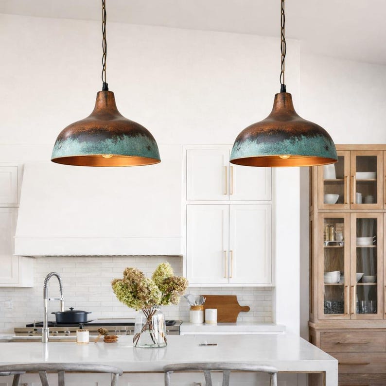 Puede incluir: Dos l&aacute;mparas colgantes con pantallas de color cobre y una banda turquesa. Las luces cuelgan de cadenas negras en un entorno de cocina. La cocina cuenta con gabinetes blancos, una encimera blanca y un jarr&oacute;n con flores.