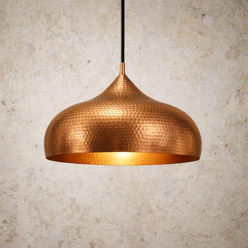 May include: A copper-colored pendant light fixture with a hammered texture. The light has a dome shape with a wide base and a narrow top, suspended by a black cord. The interior of the dome is a warm gold color.