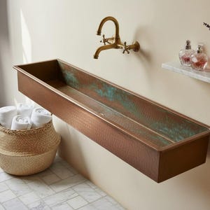 Könnte beinhalten: Ein rechteckiges Waschbecken in Kupferoptik mit Patina-Finish ist an einer beigen Wand montiert. Ein Wasserhahn aus Messing im Vintage-Stil befindet sich über dem Waschbecken. Ein geflochtener Korb enthält gerollte weiße Handtücher. Ein Marmorregal zeigt Parfümflaschen und Lavendel.