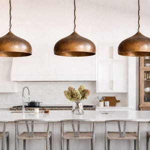 Op de afbeelding: Drie koperen hanglampen hangen boven een keukeneiland met barkrukken. De lampen hebben een gehamerde textuur en een warme, metallic afwerking. De keuken heeft witte kasten en een lichtgekleurd aanrecht.