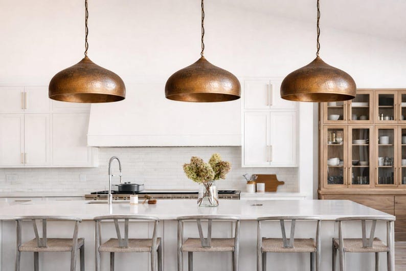 Op de afbeelding: Drie koperen hanglampen hangen boven een keukeneiland met barkrukken. De lampen hebben een gehamerde textuur en een warme, metallic afwerking. De keuken heeft witte kasten en een lichtgekleurd aanrecht.