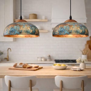 May include: Two pendant lights with a distressed blue and gold finish hang above a kitchen island. The lights have a copper-coloured top and a warm glow. The kitchen features a wooden countertop and white tile backsplash. The lights are dome-shaped.
