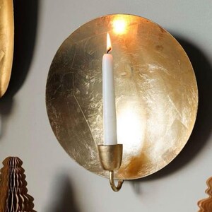 May include: A gold-coloured wall-mounted candle holder with a lit white candle. The candle holder has a circular design and a textured surface. The flame is bright and the candle is centered. Decorative items are in the foreground.