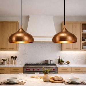 May include: Two copper-colored pendant lights hang above a kitchen island. The lights have a hammered texture and a teardrop shape. The kitchen features light wood cabinets, a white range hood, and a marble countertop. The lights are suspended by black cords.