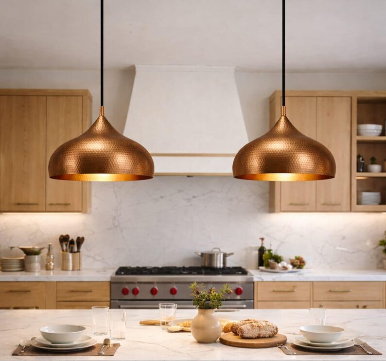 May include: Two copper-colored pendant lights hang above a kitchen island. The lights have a hammered texture and a teardrop shape. The kitchen features light wood cabinets, a white range hood, and a marble countertop. The lights are suspended by black cords.