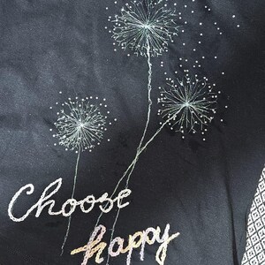 Pode incluir: T-shirt preto com design de dente-de-leão bordado e as palavras "Choose happy". Os dentes-de-leão são brancos com detalhes prateados. O texto é em branco e cores pastel.