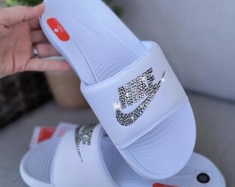 May include: White Nike slides with a rhinestone Nike logo. The slides have a textured sole and a red accent on the strap.