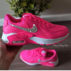 Pode incluir: Tênis Nike Air Max rosa com um swoosh branco decorado com strass. Os tênis têm uma sola branca e uma parte superior de malha rosa.