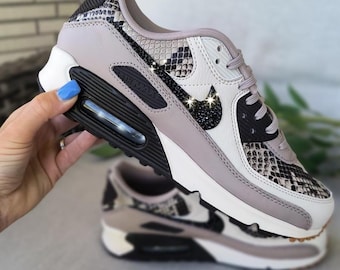 May include: A Nike Air Max sneaker featuring a snakeskin pattern on the toe and heel, light taupe leather overlays, and a black glittery Nike swoosh. The midsole is white with a black Air Max unit, and the laces are light taupe. Text reads BlingandDesignShop.