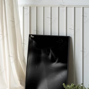 May include: Black and white framed art print. The artwork features a grayscale image of a person's upper body. The print is displayed against a white wall with vertical paneling, next to a white curtain and a small green plant.