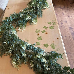 May include: A lush, artificial eucalyptus garland with varying shades of green and white leaves. The garland is draped across a light-colored wooden surface, with a subtle ivy design in the corner.