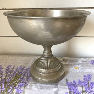 May include: A silver-toned metal pedestal bowl with a wide, smooth basin and a flared rim. The base features a decorative, ribbed column design that connects to a circular, flat foot, providing stability to the elevated vessel structure.