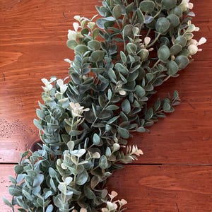 May include: A faux eucalyptus garland with green and white leaves. The garland is arranged on a wooden surface, creating a natural and decorative element. The leaves have a soft, textured appearance, adding a touch of greenery to any space.